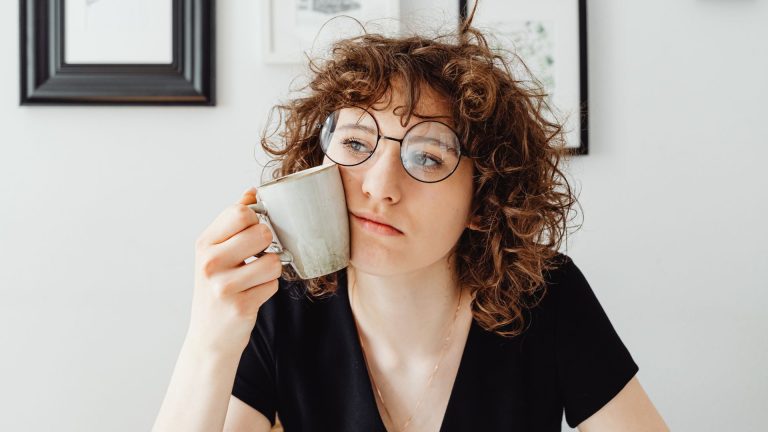 En la imagen aparece una mujer con gagas con una taza de café apoyada en su cara y mirada perdida. Es nuestra imagen del artículo de Presentismo de nuestro blog